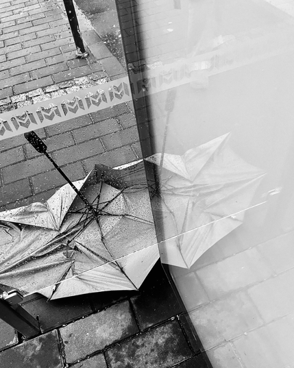 A black-and-white image of a broken umbrella lying on wet pavement, its reflection visible in a glass surface nearby.