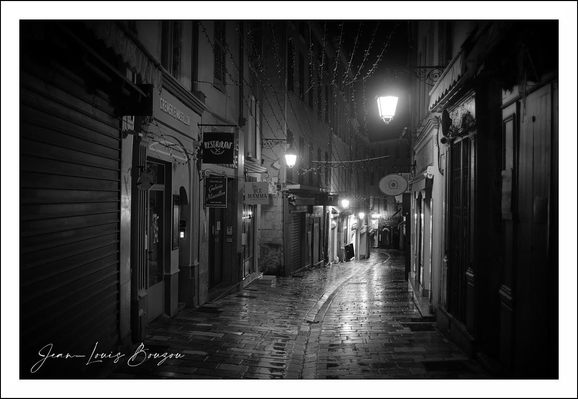 This black-and-white photograph captures a narrow, glistening street at night. The cobblestone pathway reflects the soft glow of the street lamps, creating a shimmering effect on the wet surface, likely from recent rain. Buildings closely line both sides of the alley, their facades displaying a variety of architectural details—windows, signs, and doorways—that add to the urban texture.
The street is empty except for a distant figure, who seems to be walking away, lending a sense of solitude and quiet to the scene. Overhead, subtle strands of lights are draped, possibly festive decorations, adding a delicate contrast to the otherwise stark monochrome setting.
Symbolically, narrow, dimly lit streets often evoke feelings of mystery, introspection, or nostalgia. The wet cobblestones can symbolize freshness or renewal after a storm, while the lone figure might represent solitude or a journey. Artistically, the choice to shoot in black and white emphasizes contrasts and textures, stripping away distractions of color to focus on mood and composition.
Overall, the image conveys a contemplative urban spirit, inviting viewers to imagine stories behind the empty passage and the quiet night.
