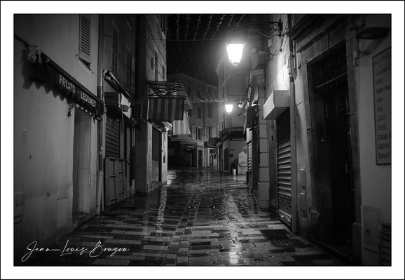 This evocative black-and-white photograph depicts a narrow, rainy street at night, creating a scene rich in atmosphere and mood. The wet cobblestone pavement reflects the dim glow of street lamps, casting shimmering pools of light that contrast with the enveloping darkness. The slick ground and the slight blur of reflections amplify the sense of quiet solitude.
The architecture lining the street carries a sense of aged charm—shuttered shop fronts, wrought iron signs, and modest awnings extend tentatively over the sidewalks. The narrowness of the street, along with the cobblestones' irregular pattern, suggests a historical or European setting, where streets are intimate and winding rather than wide and orderly.
The vintage-style lamp on the left emits a soft, haloed light, whose warmth feels intimate yet melancholic amid the absence of people. Further down the street, shadows deepen, evoking mystery and drawing the eye into the distance.
The monochrome palette emphasizes texture and contrast, lending a timeless quality to the image. Symbolically, rainy streets at night often evoke introspection, solitude, or transition—moments where the bustle of day fades and the quiet unknown emerges. Artistically, this photo captures a moment suspended between movement and stillness, light and shadow, presence and absence.
It is a contemplative tableau of urban solitude, inviting viewers to imagine the stories hidden in the empty alleys and the quiet poetry of night rain on stone.
