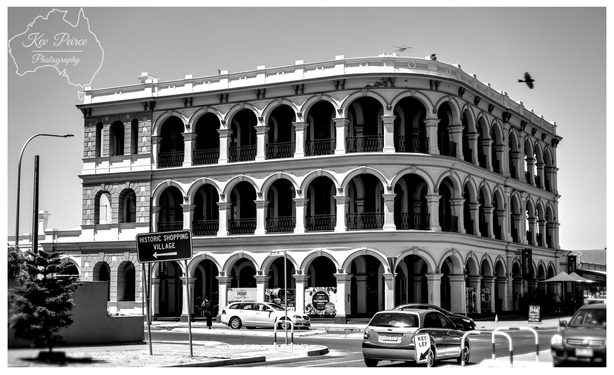 A black and white photograph of the historic three story Largs Bay Hotel in South Australia, featuring a striking facade with arched verandas on the second and third floors.

The architecture is detailed with numerous repeating arches and small balconies with balustrades.

In the foreground, there is a street corner with a few parked cars, and a traffic sign points to a "HISTORIC SHOPPING VILLAGE" to the left. A bird is visible in flight over the rooftop on the right side.