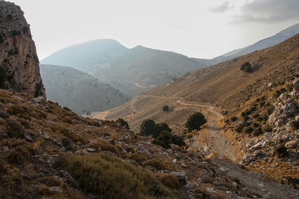 A scenic landscape featuring rolling hills and rocky terrain. A winding dirt path leads through the foreground, with sparse vegetation and scattered trees. The atmosphere is hazy, creating a soft and muted color palette.