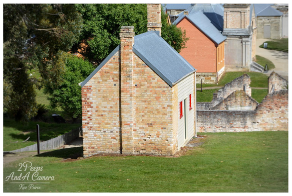 A photograph signed by Kev Peirce showing a small, one story historic brick building with a light grey corrugated iron roof and a narrow wooden side wall featuring two small windows trimmed in red.

The building is situated on a green, grassy slope at the Port Arthur historic site, with tree foliage on the left and the ruins of larger, darker brick structures visible in the blurred background to the right.