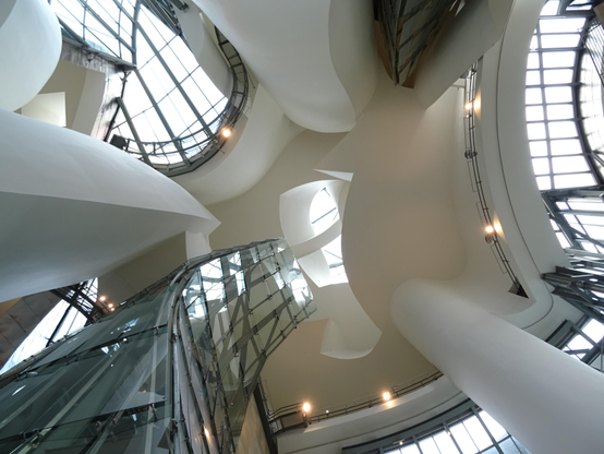 Im zentralen Raum des Guggenheim-Museums von unten nach oben über alle Ebenen aufgenommen. Man sieht Säulen, Fenster, Wände und Lichtspiele, die den Raum gewissermaßen tanzen lassen.