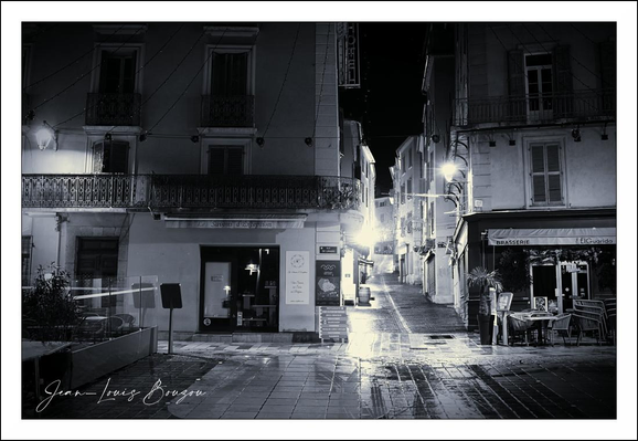 This black-and-white photograph captures a quiet, atmospheric street scene, likely in a European town known for its charming architecture. The wet pavement reflects the soft glow of street lamps, adding a shimmering texture that enhances the night-time ambiance. The buildings on either side of the narrow street showcase classic architectural elements like balconies with wrought-iron railings, shuttered windows, and a slightly worn facade that hints at history and stories untold.
To the right, an outdoor seating arrangement under an awning suggests a small café or bistro, inviting passersby to sit and absorb the town's tranquil nightlife. The absence of people intensifies a sense of stillness, almost like a frozen moment in time, allowing the viewer to imagine the quiet hum of a late evening.
Symbolically, the monochromatic tone lends a timeless quality to the scene, evoking nostalgia or contemplation. Streets like this often symbolize a journey or passage, and the light at the end of the street could metaphorically represent hope or the promise of discovery.
Overall, the photograph beautifully balances light, shadow, and texture to create a moody and evocative visual narrative.
