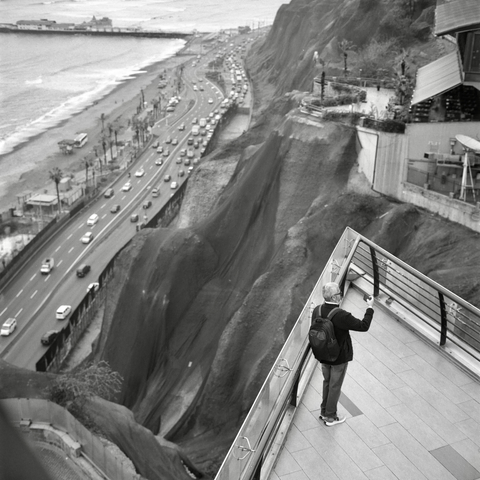A black-and-white film photo. A man on a terrace is taking a photo high above the adjacent traffic and sea in Lima, Peru. Friday, September 26, 2025.