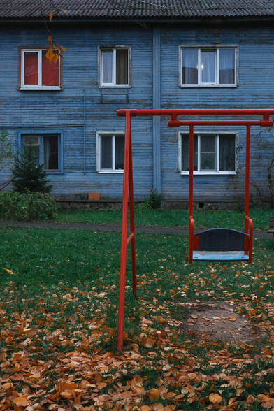 In the foreground, there is a red metal swing. In the background, there is a blue wooden two-story house with a window covered by red curtains. Between the house and the swing, there is green grass and orange, fallen leaves.