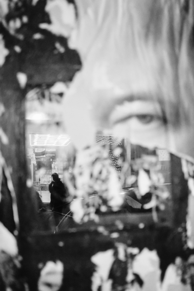 A silhouette is seen through a refection of a construction fence window previously covered with a poster of a person's face that has been torn off leaving only the top right eye and forehead of the face.