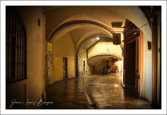 This image captures a serene and warmly lit passageway, likely in a historic European town or village. The scene is bathed in a golden glow, highlighting the rich texture of the aged stone floor and the smooth, rounded arches overhead. The architectural elements evoke a sense of timelessness—arched ceilings and narrow corridors characteristic of medieval or Renaissance structures.

The way the light filters through the tunnel space creates a layered effect, with shadowed areas gradually giving way to brighter spots near the exit of the passage, inviting the viewer’s gaze to follow the path beyond. The polished cobblestones reflect the light softly, enhancing the peaceful and almost mystical atmosphere of the setting.

The open door on the right adds a hint of narrative possibility, as if inviting a visitor to step inside and explore what lies within. The small hanging sign near that door suggests a merchant shop or a quaint café, contributing to the cultural charm of the scene.

Overall, this space feels like a quiet, almost hidden part of a town with rich history—offering a glimpse into a slower, more contemplative rhythm of life. 