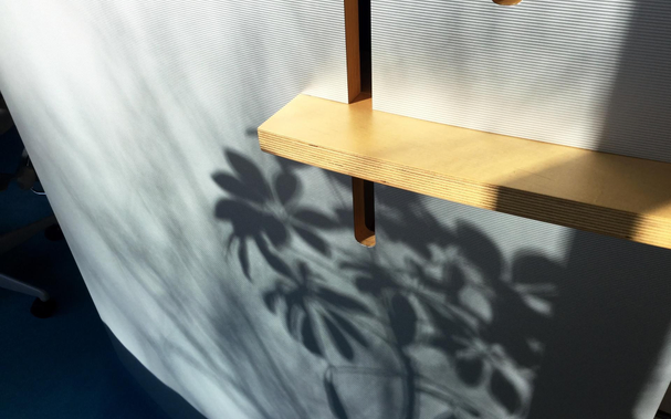 Sunlight and shadows from a plant hitting a textured vertical panel with plywood shelf of a desk at work.