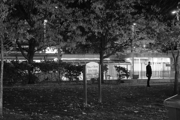A small park at night, the sign visible in mid frame. The far background is the lights of the subway station itself, the left of frame obscured by the trees in the mid ground, the right side glowing with internal light. Silhouetted against these lights, a man stands in the park smoking a cigarette.