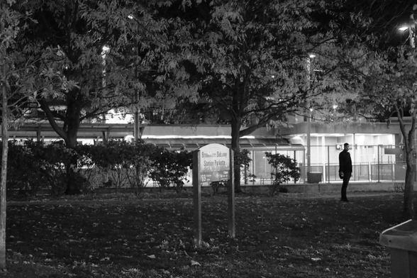 A small park at night, the sign visible in mid frame. The far background is the lights of the subway station itself, the left of frame obscured by the trees in the mid ground, the right side glowing with internal light. Silhouetted against these lights, a man stands in the park smoking a cigarette. 
