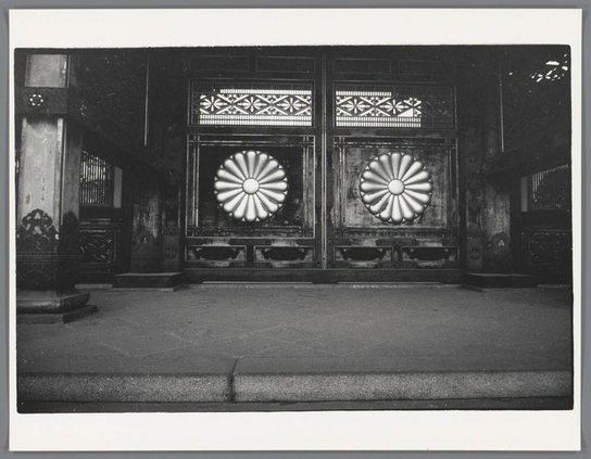 The image depicts a set of ornate doors with intricate designs. The doors are large and appear to be made from dark wood, with metal reinforcements at the corners for durability. On each door panel, there is an oversized circular pattern resembling a flower or sunburst design, which adds decorative flair while also serving as structural supports.

The upper part of the door features ornamental lattice work that includes geometric patterns such as diamonds and crosses, typical of traditional Japanese architecture known as "kagamigasa" style. This type of decoration is often found on shrine doors to symbolize protection against evil spirits.

To the left side of the image, a portion of what seems like a smaller structure or entranceway can be seen with similar decorative elements. The pavement in front appears well-worn, suggesting frequent use and possibly indicating that this location has historical significance.

The photograph is monochromatic, emphasizing textures and contrasts rather than colors. There's no discernible movement; the setting conveys stillness, perhaps evoking a sense of reverence or tradition associated with such architectural features found at temples in Japan. The absence of people adds to the serene atmosphere conveyed by this image.