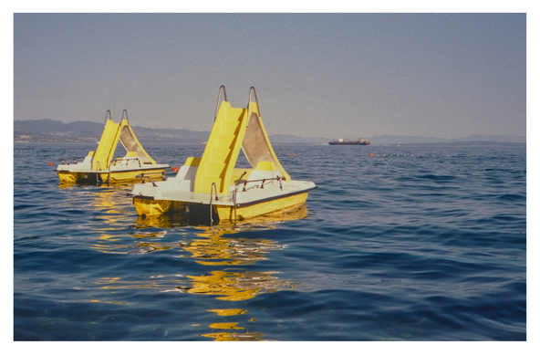Color photograph of two yellow pedal boats with slides floating in the blue water off the beach at Opatija. A container ship can be seen further out at sea in the background.