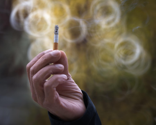 Photo of a hand holding up a cigarette. The actual smoke from the cigarette is faintly visible. The background is composed of rings of light, which themselves resemble smoke.