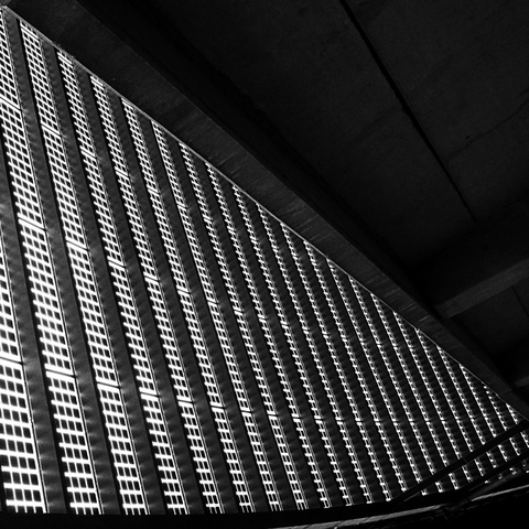 Ein Wand die schräg nach unten führt. Sie besteht aus dicken Stahlstreben die senkrecht nach unten führen. Zwischen diesen Stahlstreben ist ein Netz aus quadratischen Feldern die auch von feineren Stahlstreben gebildet werden. Das Foto ist in Schwarzweiß und am Weserstadion in Bremen entstanden. 