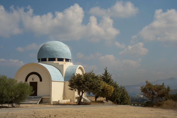 A small chapel featuring a blue dome and unique rounded architecture, surrounded by dry landscape and trees, under a partly cloudy sky.