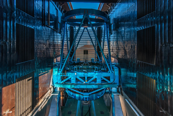 A blue toned photography of the Subaru Telescope enclosure opened in preparation for observations. Beyond the slit, Maunakea can be seen illuminated by the setting sun. 