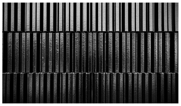 Das Bild zeigt eine streng geometrische, schwarz-weiße Struktur aus vertikalen, stark kontrastierenden Streifen. Die Oberfläche wirkt hart und leicht glänzend, mit einer unregelmäßigen, leicht körnigen Textur. Die Streifen sind in mehreren horizontalen Segmenten angeordnet, die durch feine Linien voneinander getrennt sind. Insgesamt entsteht ein sehr rhythmisches, fast industriell wirkendes Muster.

Wer nicht so ganz versteht was das ist: das ist eine Art keramische Beplankung an der Außenwand eines Gebäudes. ;-)