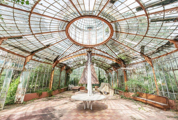 An abandoned glasshouse with overgrown plants, featuring a circular skylight and a central white table surrounded by neglected furniture.