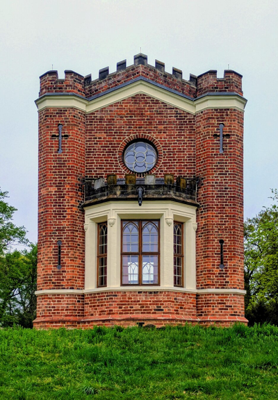 A striking red-brick tower, reminiscent of a small castle or turret, set against a backdrop of lush greenery. The structure features a cylindrical design with a crenelated parapet at the top, adding a medieval aesthetic. A prominent circular window, adorned with decorative glass panes, sits above a projecting bay window with three arched sections. The bay window is framed by cream-coloured brickwork, providing a pleasing contrast to the red brick. The tower is surrounded by a well-maintained grassy area, with trees visible in the background, enhancing the picturesque and timeless charm of the scene.