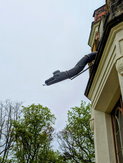 A whimsical architectural detail of a building exterior, featuring a decorative rainwater downspout shaped like a stylised dragon or snake. The snake’s head, with its open mouth and pronounced teeth, extends outward from the building’s cream-coloured façade, appearing as if it is spouting water. The building itself showcases ornate detailing, including arched windows and decorative mouldings. In the background, leafy green trees rise against a pale, overcast sky, adding a natural contrast to the building’s intricate design. The scene combines elements of fantasy and traditional architecture, creating a unique and charming visual.
