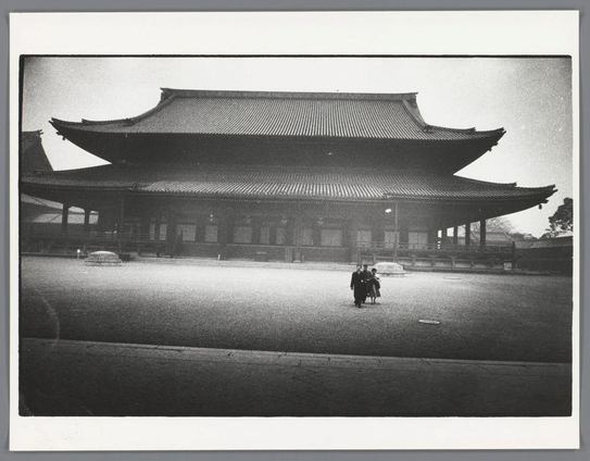 The image shows a black and white photograph of a traditional East Asian temple with distinct curved eaves typical of such architecture. The structure appears to be made primarily from wood, featuring multiple tiers in its roof design that are characteristic of historical buildings like temples or palaces found across countries including China, Japan, Korea, Vietnam, Cambodia, Laos, and Myanmar. In the foreground, there's an open paved area leading up to a large temple entrance with several individuals walking towards it; two adults appear to be guiding one child on their shoulders. The people are dressed in dark clothing which suggests this photo could have been taken during cooler weather or as part of cultural dress for visiting such sites.

The composition is simple, focusing on the contrast between the architecture and its environment. There's no visible vegetation around, indicating that this temple might be situated within an urban setting rather than a rural one where nature often surrounds historic buildings. The absence of modern elements in clothing or technology implies historical significance to the photograph, potentially from early 20th-century Japanese photography as suggested by your reference.

The image is presented on what appears to be a photographic paper border and shows signs of wear like creases at its corners which could indicate it's been handled multiple times. This gives an impression that th [...]