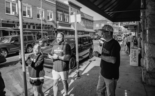 Three people on a sidewalk of a small town.  A couple on the left look to be taking photos with their cell phones.