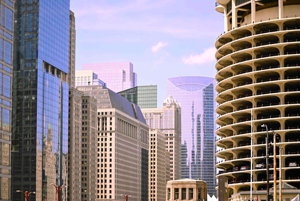 A tight photo of many skyscrapers, including some contemporary fully-glazed towers, some older towers which are mostly stone or concrete, and a tall, cylindrical, open-air parkade on the right side.