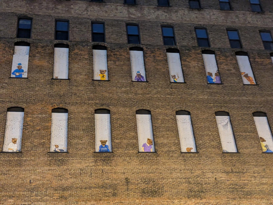 Photo of several stories of the side of a brown brick building with tall, rectangular window cutouts. Two floors of windows have murals of bears dressed and acting like people painted in the cutouts. Above those floors are real windows.