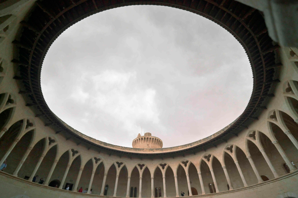 A view looking up from inside a circular courtyard in a Gothic-style Castle toward a cloudy sky, with the castle tower peeking above the far edge of the courtyard.