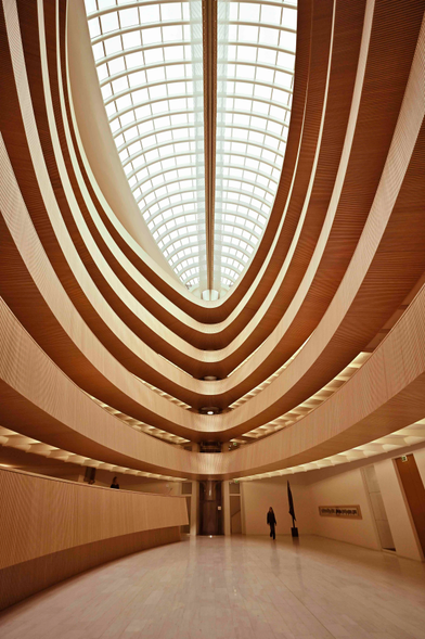 A view up toward a massive elongated, ovoid skylight, bisected by a wooden beam, framed by somewhat off-centre rings of wood panels for each floor of the atrium. The floor below is a glossy, polished, light hardwood. The sky through the skylight is cloudy.