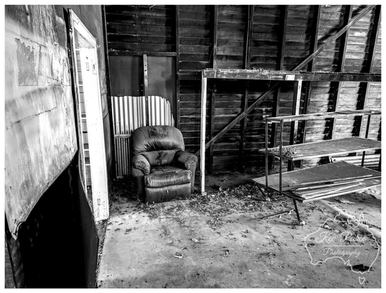 A black and white photograph of a dark, worn armchair sitting alone on a dusty concrete floor inside a decaying, abandoned wooden shed or workshop.  To the right is a rusty metal shelving unit. An open doorway on the left provides a bright contrast. The image is signed Kev Peirce.