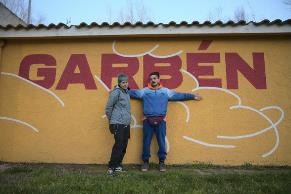 Photo of two people posing in front of a building painted in ochre with some large text painted in red over it. The text spells "Garbèn", the local name for south wind.
