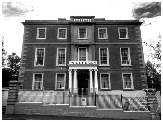A powerful, straight on black and white photograph of Westella House, a three-story, symmetrical historic building with a central portico featuring columns.  The name 'WESTELLA' is visible above the main entrance. The foreground includes a low wall and black metal fencing.