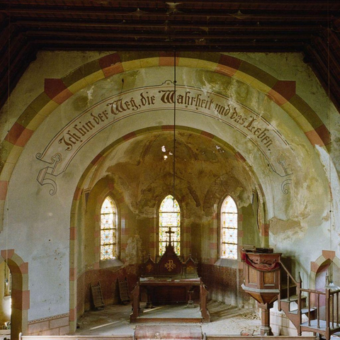 An abandoned church interior with faded walls, arched windows, a wooden pulpit, and German text above the altar.