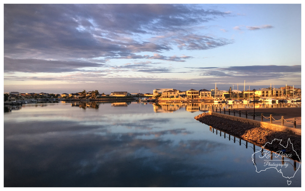 A wide angle landscape photograph of the Wallaroo Marina at sunset. The still water of the marina reflects the cloudy sky and the luxury homes lining the waterfront.  Several sailing boats are moored on the right, next to a rock retaining wall and a fenced path bathed in golden light. The overall mood is peaceful and serene.