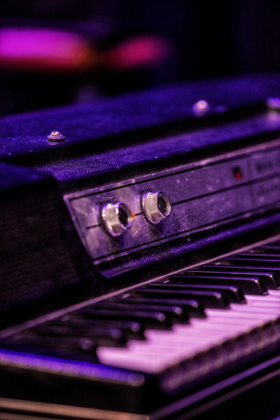 Auf dem Bild ist eine alte elektronische Tastatur von einem Wurlitzer E-Piano  abgebildet. Das Instrument weist schwarze und weiße Tasten sowie ein Bedienfeld mit zwei silbernen Drehknöpfen und einem roten Licht auf. 
Der untere Teil des Bildes zeigt die Klaviatur mit den typischen schwarzen und weißen Tasten.
Der obere Teil des Instruments verfügt über ein schwarzes Bedienfeld mit zwei großen, runden, silbernen Reglern.
Das Instrument ist in warmes, violettes und pinkes Licht getaucht, was auf eine Bühnen- oder Studioatmosphäre hindeutet.