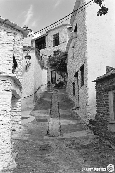 Narrow, steep cobbled alley in Capileira, lined with whitewashed stone buildings, barred windows, and a vintage street lamp; the street curves uphill under a mostly clear sky.