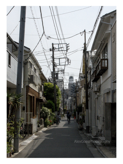 A small street in Tokyo. Two stories houses lines up in shades of greys, with a hint of green from plants, with higher buildings in the distance. Electric cables crisscross the sky. Two people on bycicles are riding away from the camera. The sky is grey.