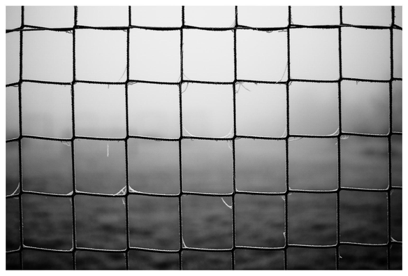 Das Bild zeigt ein engmaschiges Draht- oder Kunststoffnetz, vermutlich ein Zaun oder ein Sportnetz. Die Aufnahme ist in Schwarzweiß gehalten und wirkt durch den Nebel im Hintergrund sehr atmosphärisch.

Das Netz steht im Vordergrund scharf im Fokus; an einigen Knotenpunkten hängen kleine Reste von Spinnweben oder feinen Fäden. Der Hintergrund ist stark verschwommen und kaum erkennbar, was die Struktur des Netzes noch stärker betont. Insgesamt vermittelt das Foto eine ruhige, leicht melancholische Stimmung.