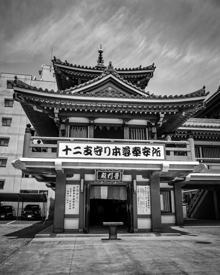 Tiene el aspecto de un templo de 2 plantas, con vigas de madera, paneles blancos en las paredes y techos curvados y con tallas muy elaboradas. Como los típicos templos japoneses. Este, sin embargo, es un pequeño anexo del templo principal donde, creo, vendían souvenirs del templo (amuletos, principalmente). Hay un cartel grande con símbolos japoneses que dice algo sobre los dioses guardianes de los símbolos del zodiaco. Blanco y negro.