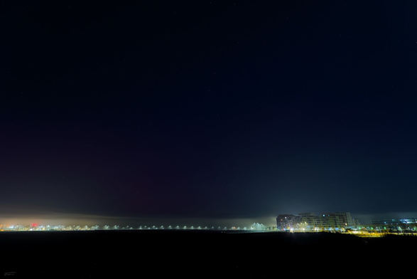 Das Foto zeigt eine nächtliche Szene mit einem weiten, dunklen Himmel, unter dem eine gerade Reihe von Straßenlaternen und beleuchteten Gebäuden am Horizont zu sehen sind. Auf der rechten Seite sind mehrere größere, beleuchtete Gebäude auszumachen, während links und mittig die Lichter in einer Linie angeordnet erscheinen. Der Himmel nimmt einen Großteil des Bildes ein, dominiert durch Dunkelheit und vereinzelt schwache Sterne, während die Häuser und Laternen mit ihrem Licht und Schein im aufziehenden Nebel einen klaren Kontrast bieten.