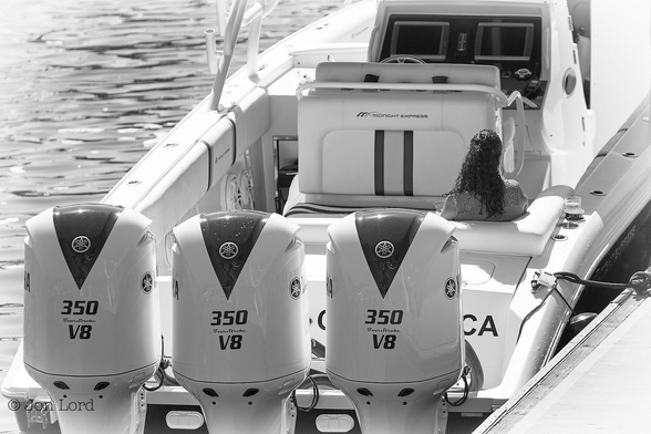 This is a black and white photo in landscape format of the rear of an open top, moored, power boat with a woman sitting in the stern. Long Beach, California (2014).

Resting on the base of the image are three very large outboard motors attached to the transom of this moored power boat. The motors are all white except for an inverted black 'V' on the top. In bold black numerals are the numbers 350 and V8, the 350 I'm guessing is the horse power. Forward of the three engines is a woman sitting on the right side and facing away from the camera. She has dark hair and is holding a drink in her left hand with a further drink in a recessed holder on her right. Forward of the woman is the back of the driver's white bench seat, with the words 'Midnight Express' contrasting in black lettering. The topside of the boat is all white while the hull is black. The forward part of the boat is off camera at the top of the photo.