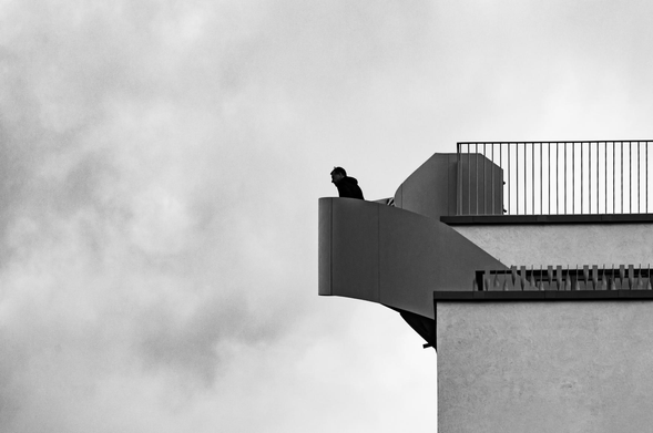 The image is a black and white photograph featuring a modern architectural structure and a silhouetted figure. The dominant element is a curved, cantilevered section of a building, appearing to be a balcony or a viewing platform. The structure is painted in shades of gray, with a smooth, almost minimalist aesthetic.

A person is standing on the edge of the cantilevered platform, their form silhouetted against the sky. Their posture suggests they are looking out or down. The silhouette is detailed enough to discern a head and shoulders, perhaps wearing a hood. The rest of the body is obscured by the darkness.