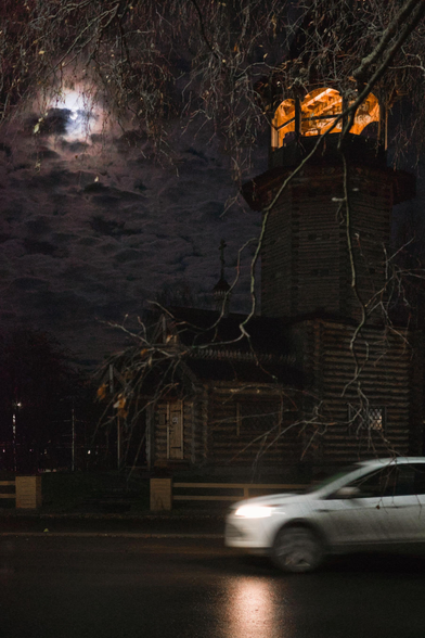 Late evening. The photo shows a wooden Orthodox church with a full moon shining next to it. The church's bell tower is illuminated. A car passes by. Tree branches are visible in the foreground.