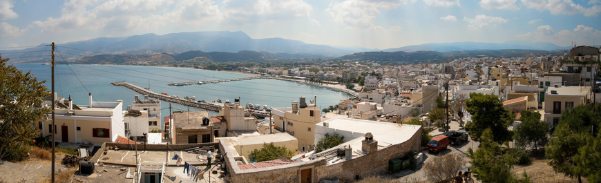 A panoramic view of a coastal town features buildings, a marina with boats, and surrounding mountains under a partly cloudy sky. The foreground includes rooftops and greenery of a Mediterranean landscape.