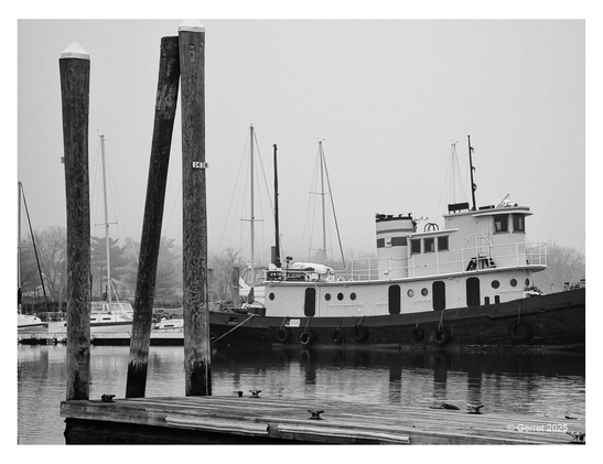 Black-and-white photo of a docked tugboat with tall masts visible in the background. Foreground has wooden dock pillars. Scene feels serene and misty.