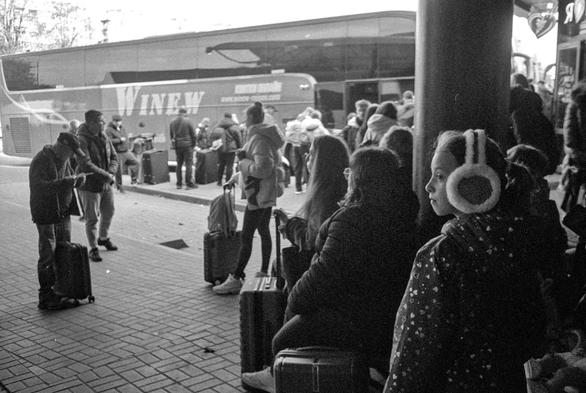 A busy bus station scene unfolds in grainy black-and-white. Travellers stand with their suitcases, waiting or checking their phones, while a large coach dominates the background. In the foreground, a girl in fluffy earmuffs looks toward the light, her profile softly illuminated against the darker crowd. The whole scene feels transient and expectant, capturing the quiet tension of departure.