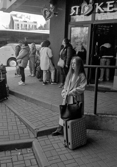 At a bus terminal entrance, a young woman stands beside her textured suitcase, holding a bag close to her. She is illuminated by soft daylight while the crowd behind her waits near a parked coach. Her long hair and calm, slightly distant expression contrast with the surrounding motion, giving the image a quiet, introspective mood.