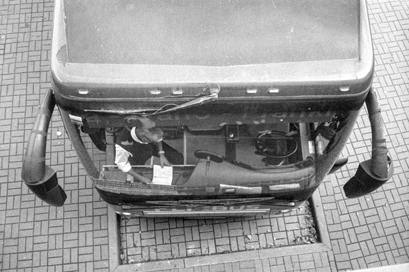 The image shows a black-and-white overhead view of a bus parked on a patterned pavement. The photo captures the bus from above, with the front of the bus visible at the bottom of the frame. Inside the bus, a person is seated, leaning forward and holding some papers in their hand. The bus's dashboard, steering wheel, and front windows are also visible. The overall scene has a gritty, high-contrast texture characteristic of analog black-and-white photography, and the perspective emphasizes the geometric lines of the pavement and the structure of the bus